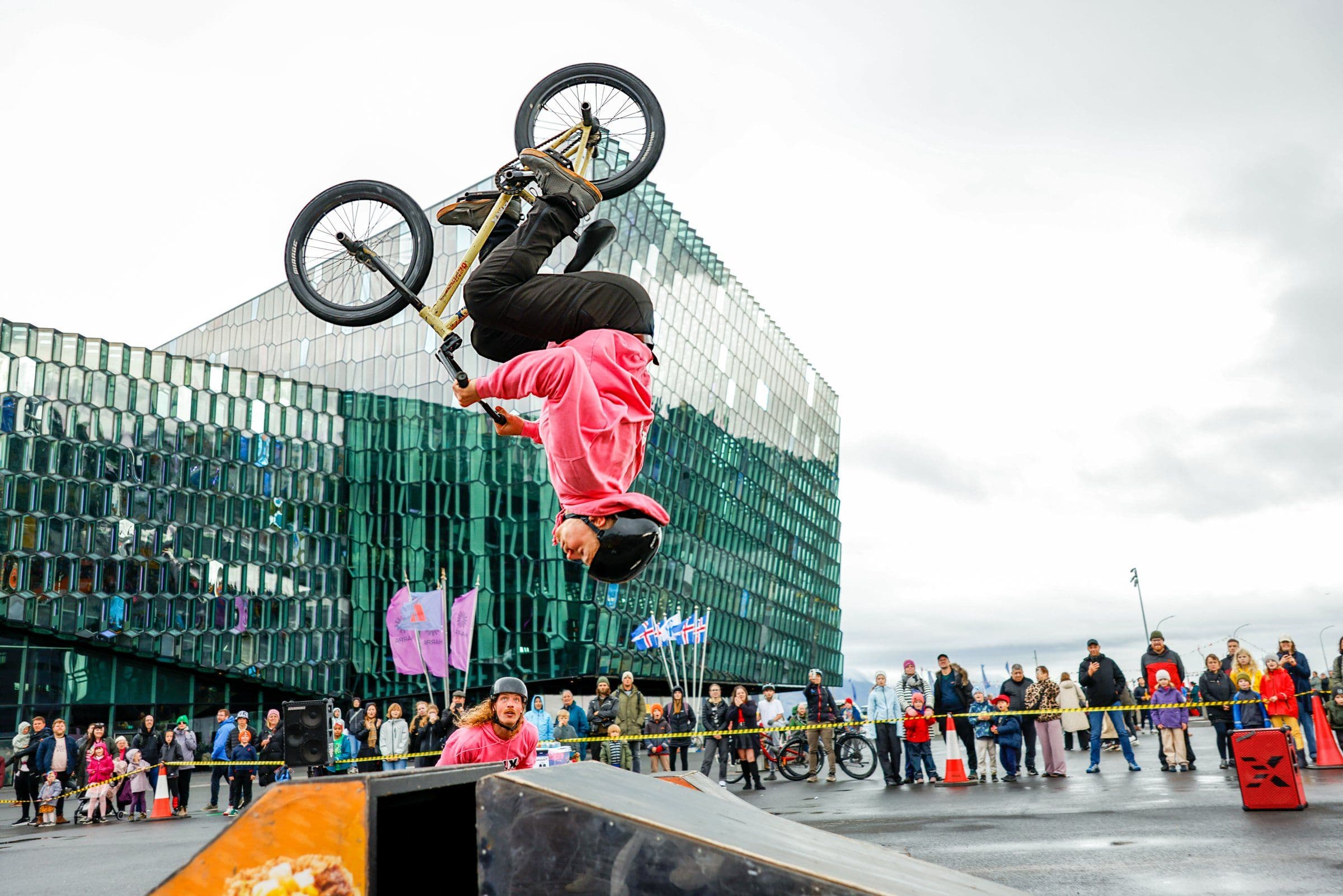 A BMX rider in a pink hoodie and black helmet performs a mid-air backflip over a ramp, with a modern glass building and a crowd watching in the background.