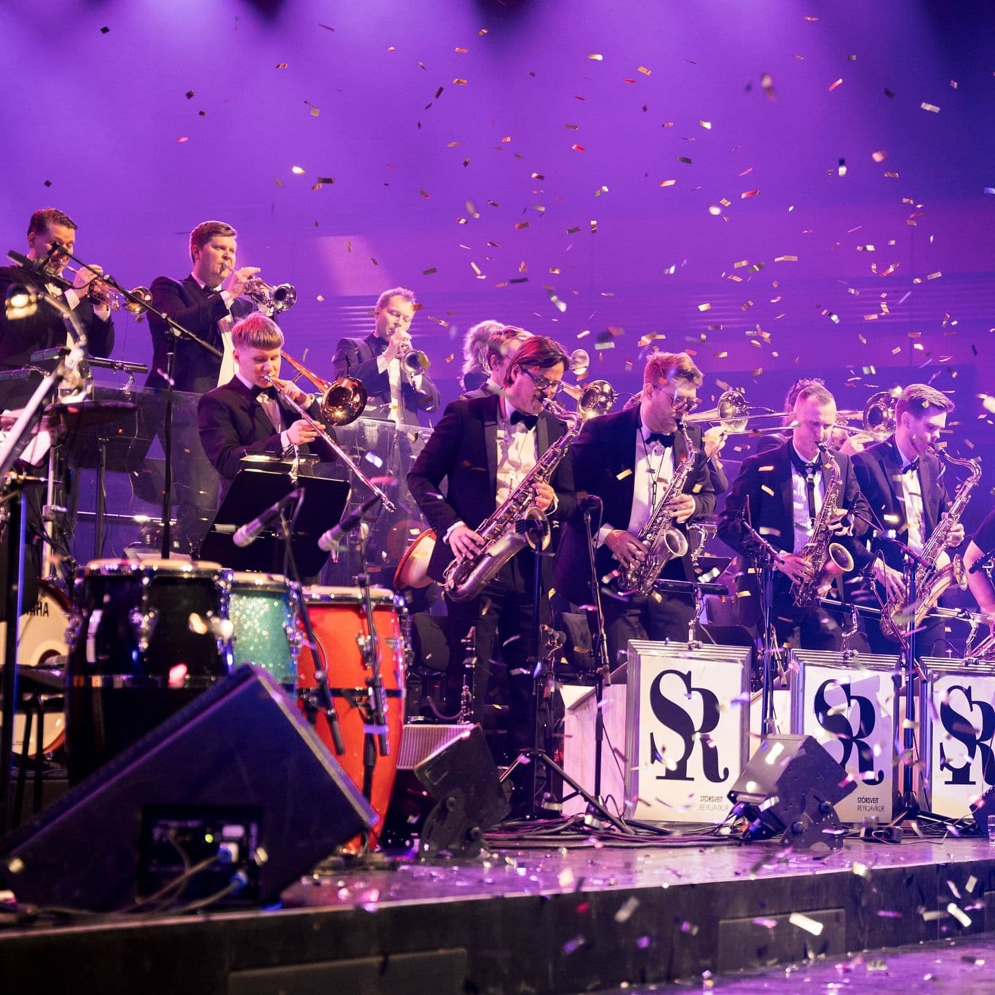 A big band in tuxedos plays brass and woodwind instruments on a stage under purple lights with confetti falling.
